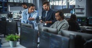 a group of men standing in front of computer monitors in a room with a plant in the middle of the room