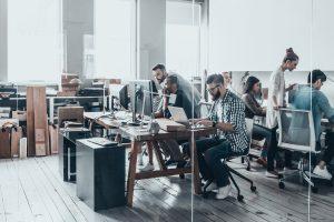 a group of people sitting at desks working on laptops in a room with wooden floors and windows