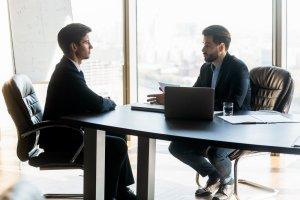 two men sitting at a table in front of a laptop computer in an office setting with a view of the city outside the window