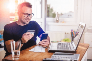 a man sitting at a table with a laptop and a cell phone and credit card in his hands and a glass of water in front of him