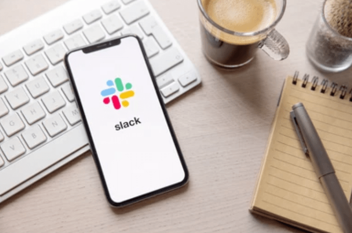 a cell phone sitting on top of a desk with slack logo, next to a keyboard and a cup of coffee next to a cup of coffee