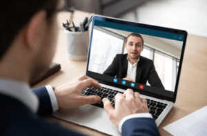 a man sitting at a desk with a laptop on his lap and a video conference on the screen behind him